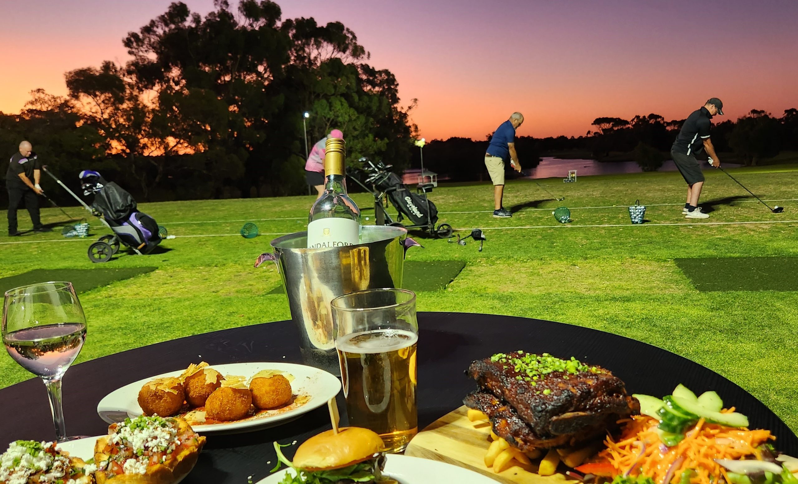 Vibrant-food-with-a-wine-bucket-beer-and-wine-and-4-golfers-in-the-background-with-a-purple-and-orange-sunset-above-the-lake